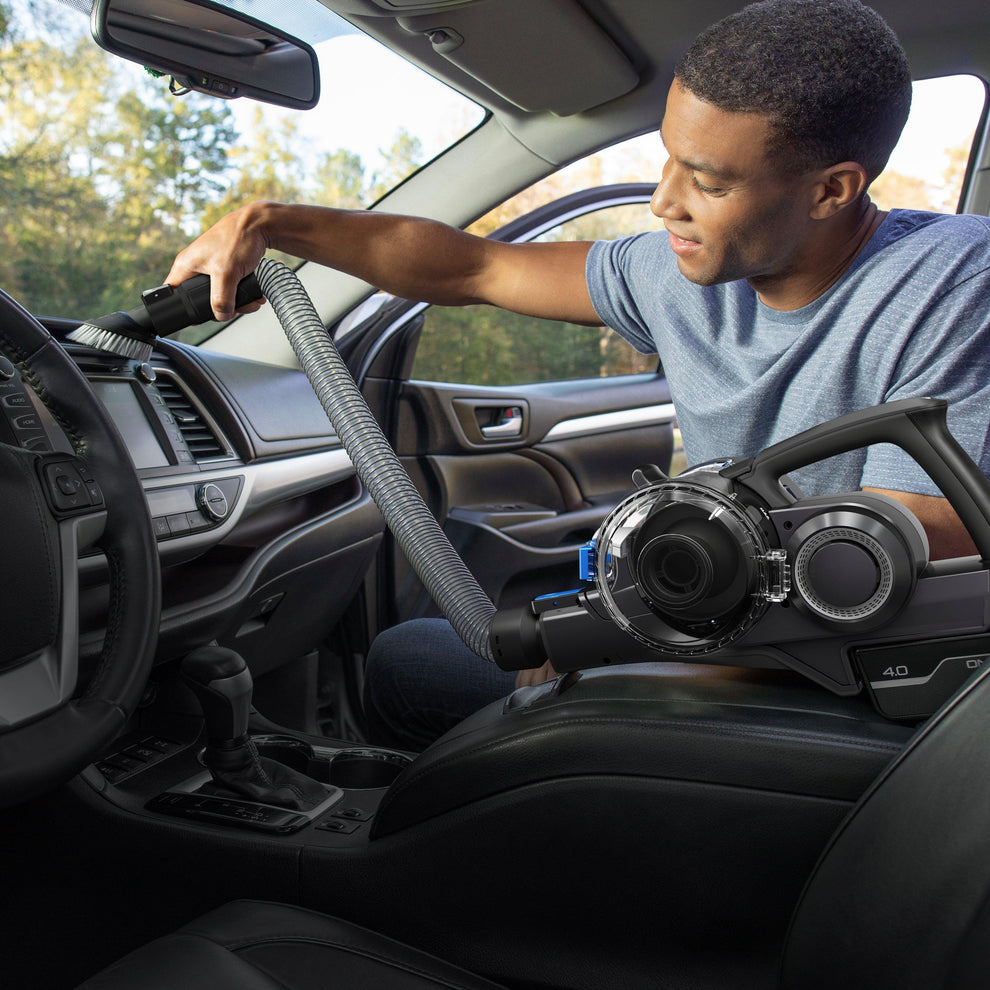 Man using a handheld cordless vacuum cleaner with an extension hose attachment to clean the dashboard area of a car's interior, highlighting the vacuum's flexibility and reach for detailed cleaning.