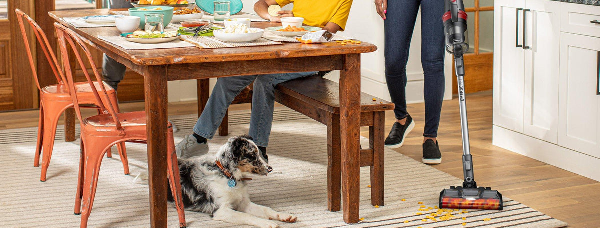 A person using their Hoover vacuum cleaner to clean up spilled crumbs and debris from the dining room floor as a dog watches.