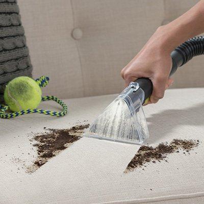 Person using a carpet cleaner attachment to remove dirt and stains from a couch, with a tennis ball nearby, showcasing effective cleaning in a pet-friendly environment.