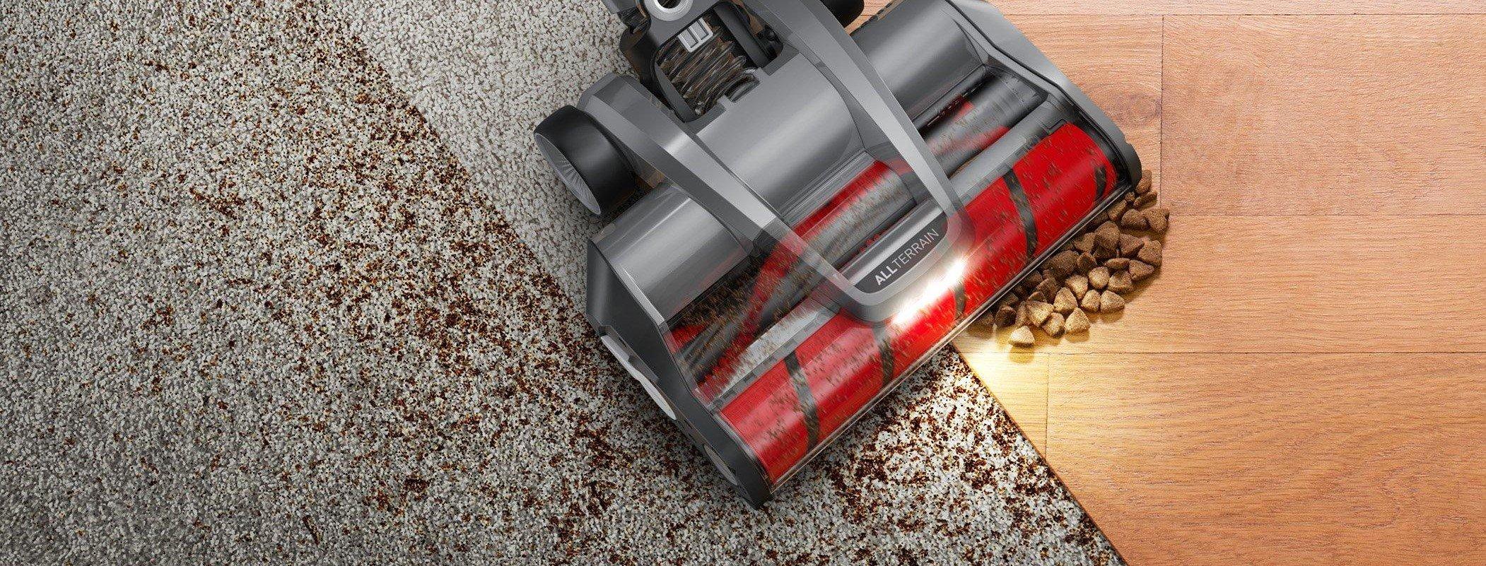 Close up view of a vacuum cleaner's brush roll head, showing its transition from carpet to hard floor, efficiently picking up debris like pet food and dirt.