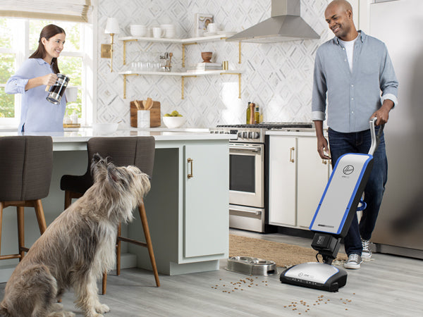 Man vacuuming dog food off of hardwood floor with woman and dog in background