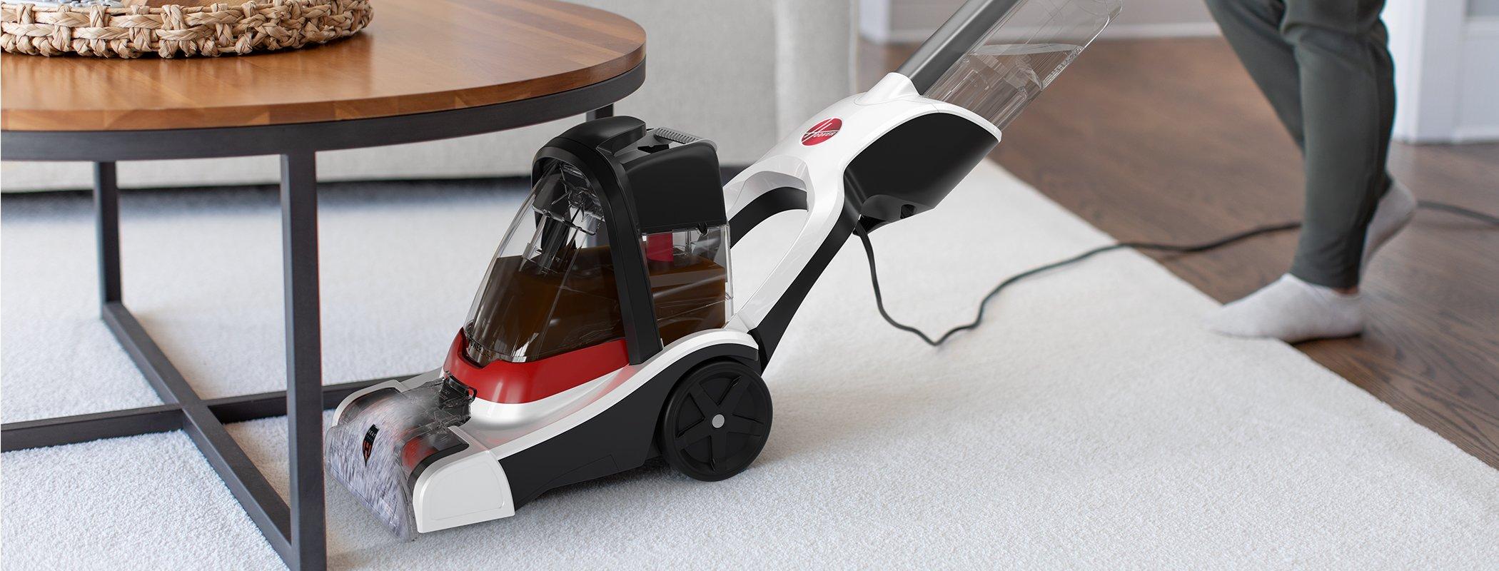 A Hoover carpet cleaner positioned near a low wooden coffee table, actively cleaning a spill on a white carpet.