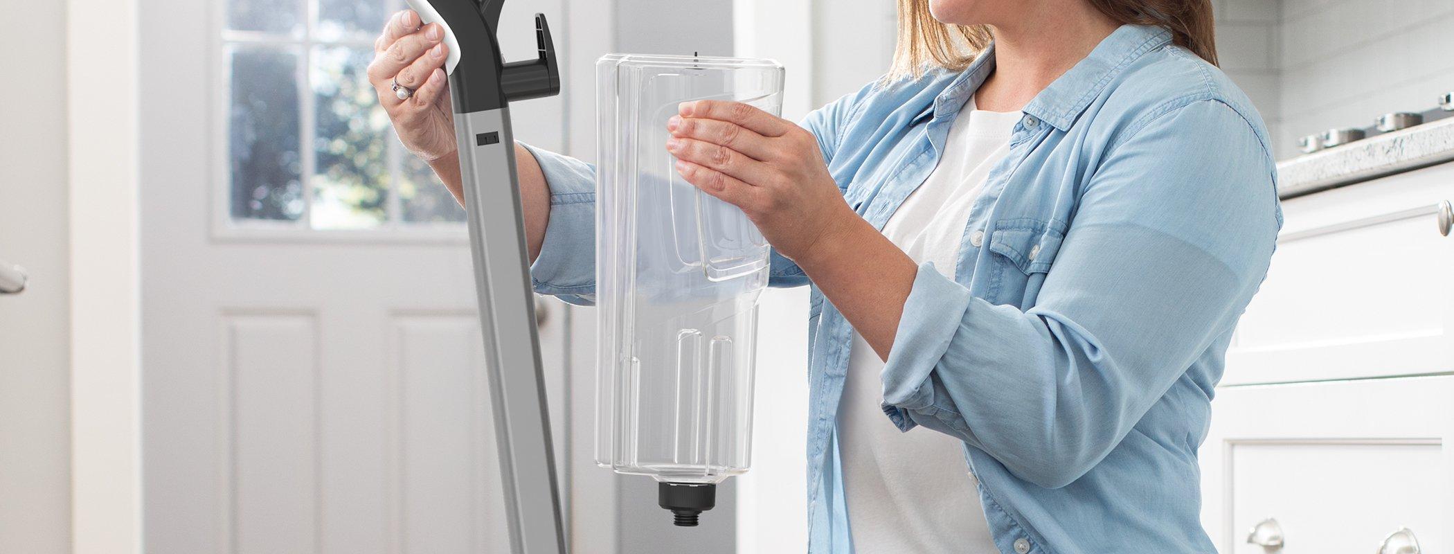 "A woman holding a transparent clean water tank for a Hoover carpet cleaner, preparing it for use in a bright kitchen setting."