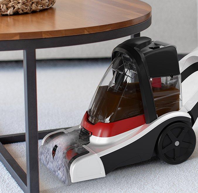 Hoover carpet cleaner positioned under a coffee table, actively cleaning the carpet, showcasing its design for effective cleaning in tight spaces.