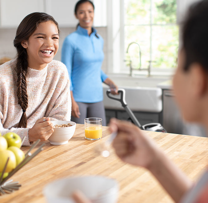 A happy family is eating in the kitchen, with a mother using her Hoover vacuum cleaner to vacuum in the background.