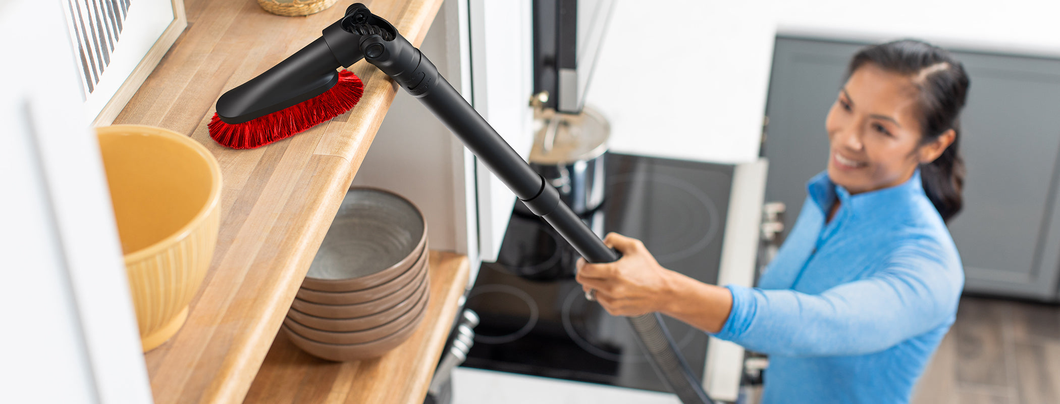 Woman using her Hoover 10-inch telescopic extension wand and dusting brush attachment to clean a hard-to-reach shelf in her kitchen