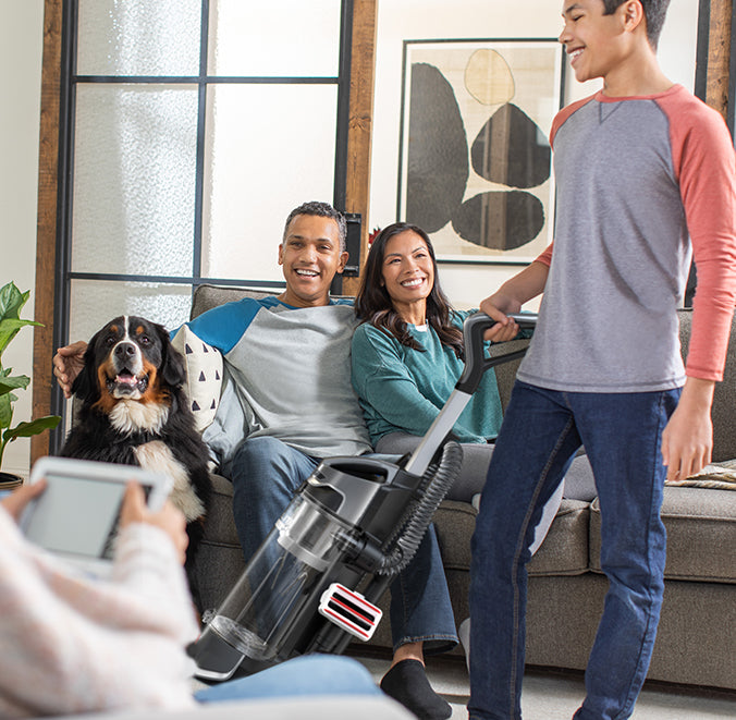 A happy family sitting on a sofa with their dog, highlighting the Hoover vacuum cleaner's pet odor prevention feature