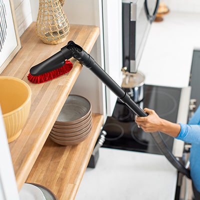 Woman using her Hoover 10-inch telescopic extension wand and dusting brush attachment to clean a hard-to-reach shelf in her kitchen