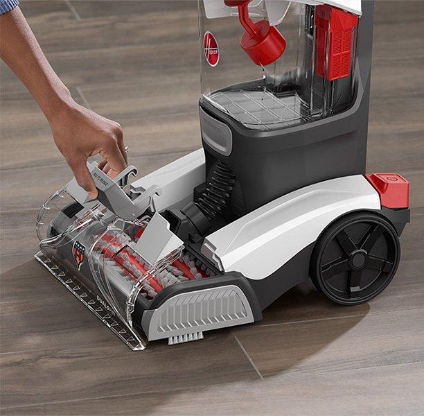 Person handling the brush roll assembly of a Hoover carpet cleaner, demonstrating ease of use and maintenance on a hard floor surface.