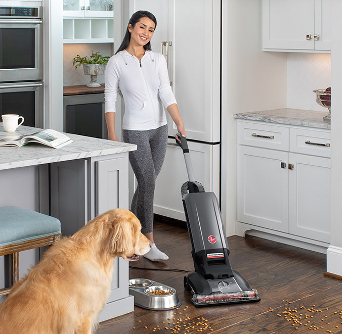 Woman using a Hoover vacuum cleaner in a kitchen to clean up scattered pet food while her golden retriever watches.