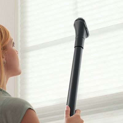 Person using a Hoover vacuum cleaner with an extended reach wand and dusting brush attachment to clean window blinds, highlighting high reach and versatility