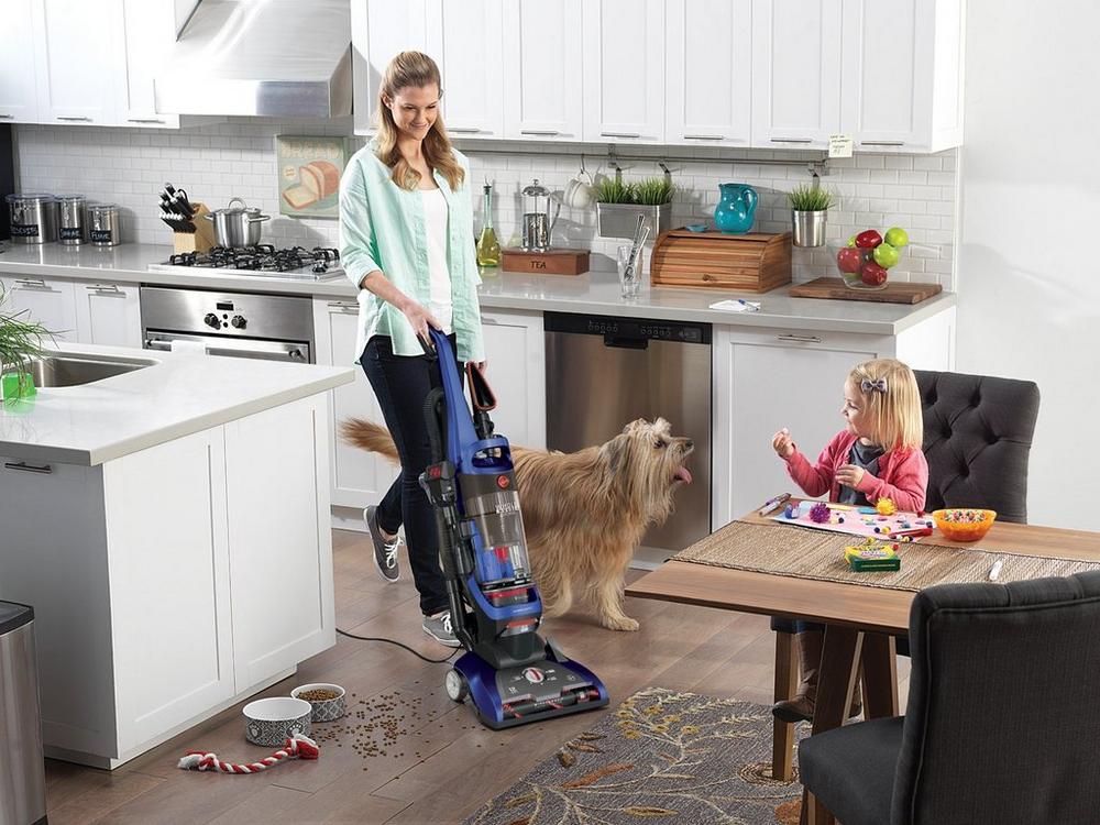 Woman using her Hoover vacuum cleaner to vacuum scattered pet food from hard wood floors in the kitchen.