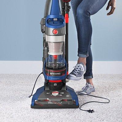 Hoover whole house rewind upright sitting on a white carpet, as someone's foot presses the retractable cord pedal for automatic cord rewind.