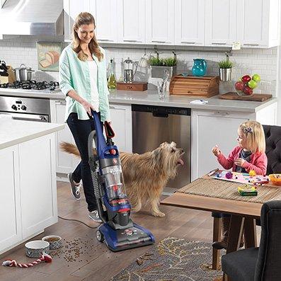Woman using her Hoover vacuum cleaner to vacuum scattered pet food from hard wood floors in the kitchen.
