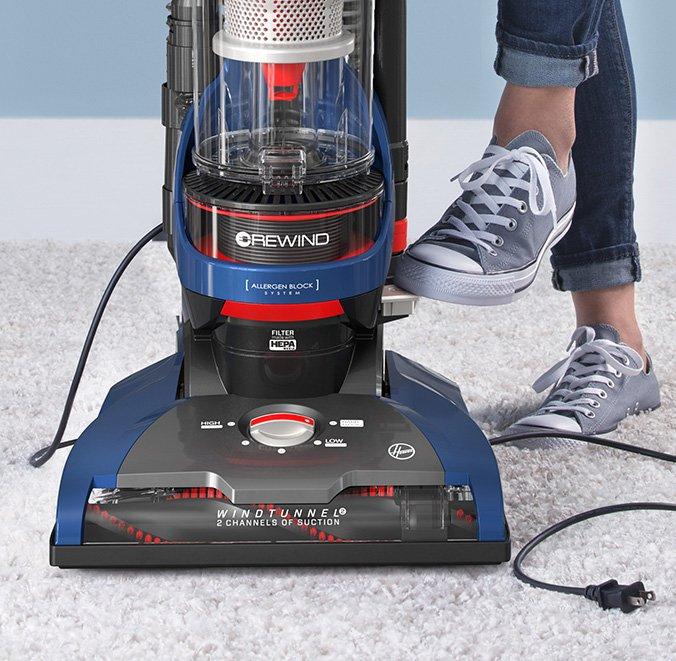 Close up of the Hoover whole house rewind upright sitting on a white carpet, as someone's foot presses the retractable cord pedal for automatic cord rewind.