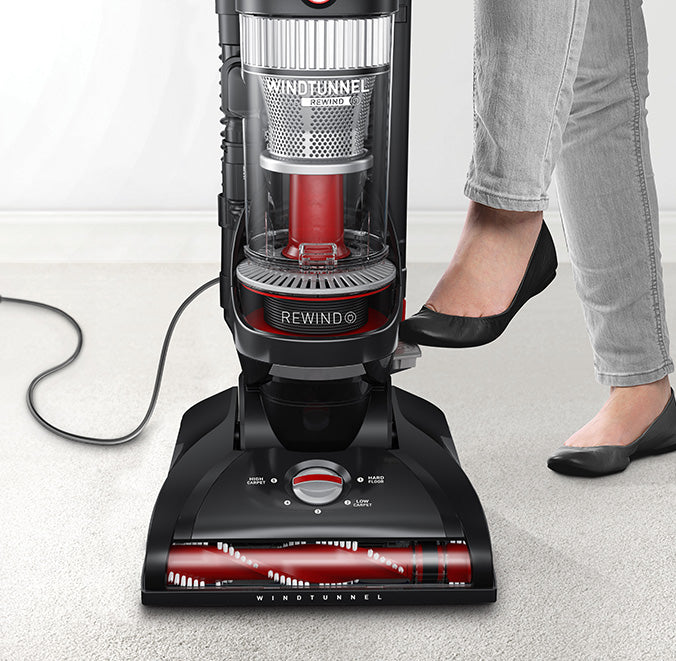 Hoover windtunnel vacuum cleaner being used on a carpet, featuring 25ft automatic cord rewind, shown by a person pressing the pedal with their foot for easy and efficient clean up