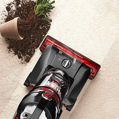 Top view of the Hoover windtunnel vacuum cleaner in action, cleaning up spilled soil from an overturned potted plant on a carpet.