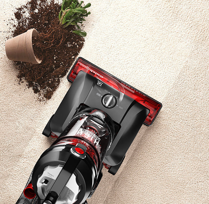 Top view of the Hoover windtunnel vacuum cleaner in action, cleaning up spilled soil from an overturned potted plant on a carpet.