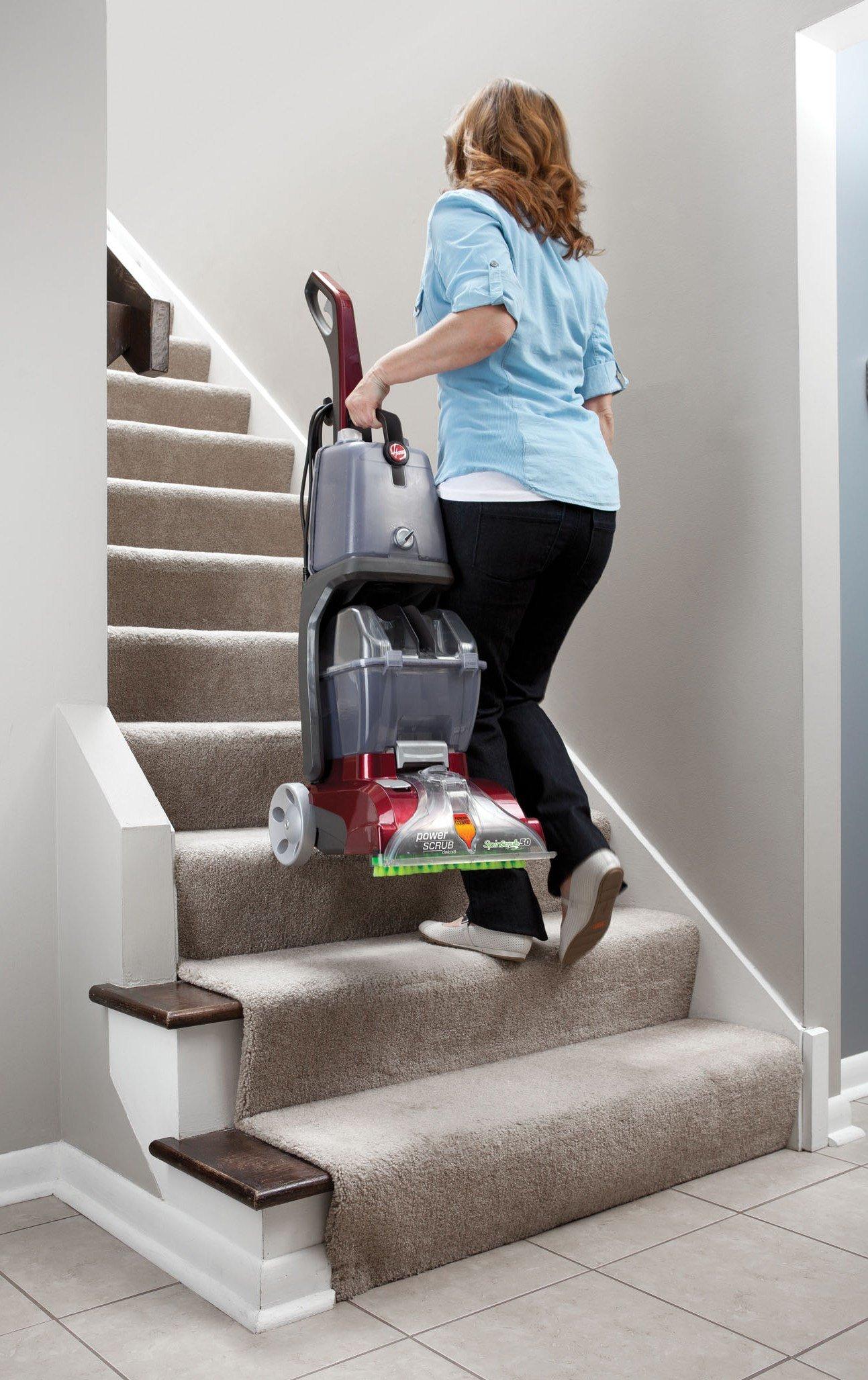 Person carrying a Hoover power scrub carpet cleaner up a flight of carpeted stairs, showcasing the lightweight and portable design