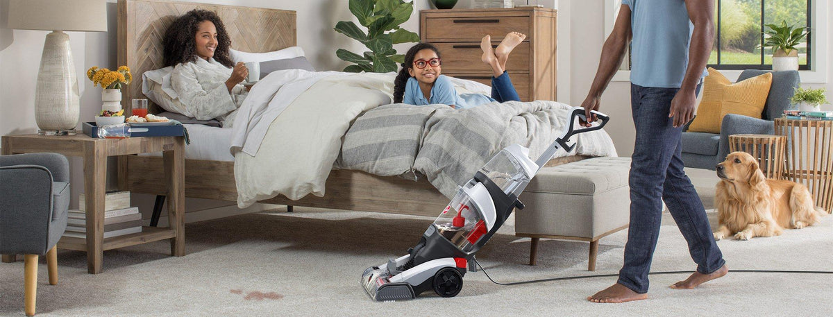 Man using a Hoover carpet cleaner in a bedroom with a woman relaxing in bed, a young girl sitting up, and a golden retriever nearby, showcasing effective cleaning in a family-friendly environment.