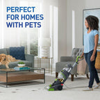 A woman using a carpet cleaner in a living room with a golden retriever lying on the couch, showcasing a well-decorated space with large windows and a coffee table.