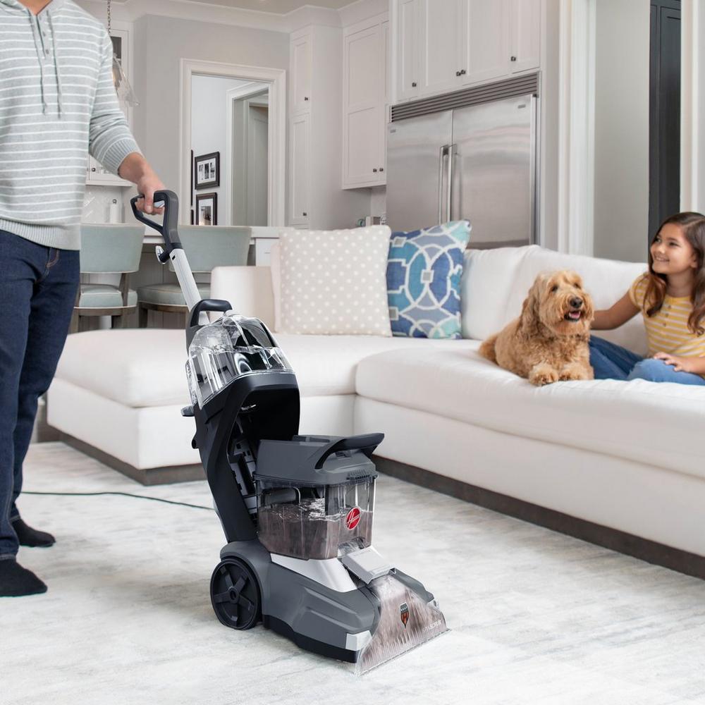 Person using a Hoover carpet cleaner on a light-colored carpet in a living room, with a dog and a child sitting on the couch, illustrating family-friendly cleaning solutions.