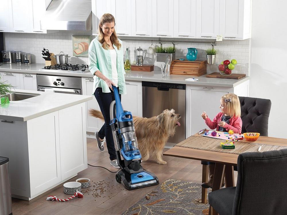 Woman using a Hoover Elite Rewind vacuum cleaner to clean a kitchen floor while a dog and child are there, highlighting effective cleaning of pet and child messes in a household environment.  