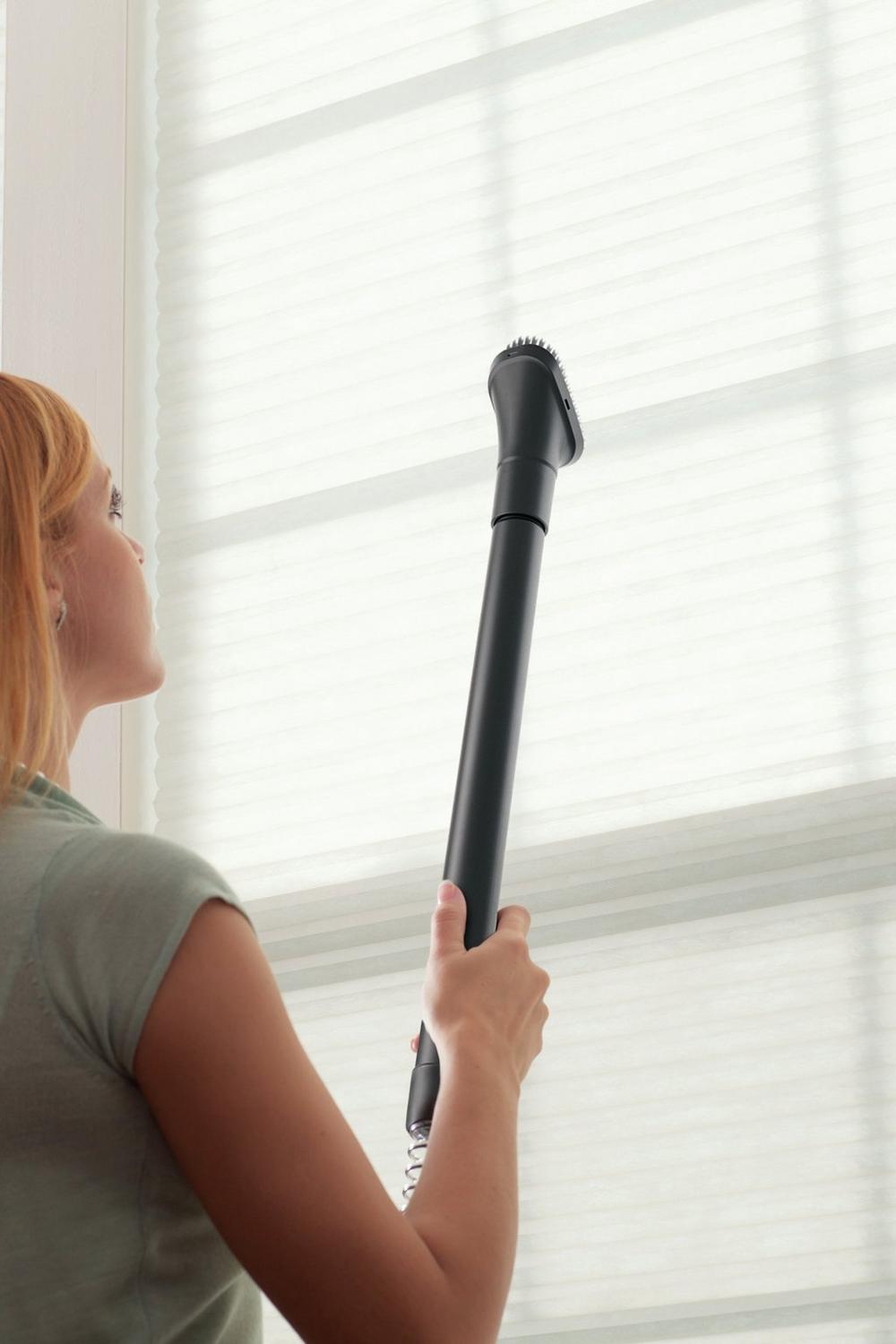 Person using a Hoover vacuum cleaner with an extended reach wand and dusting brush attachment to clean window blinds, highlighting high reach and versatility 