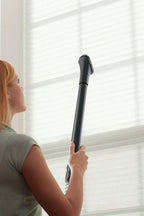 Person using a Hoover vacuum cleaner with an extended reach wand and dusting brush attachment to clean window blinds, highlighting high reach and versatility 