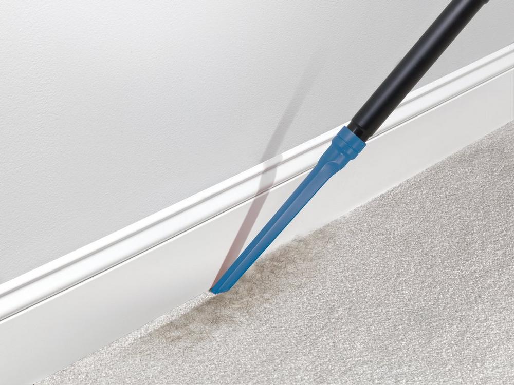 Using a Hoover crevice tool to clean along the baseboard and carpet edge, highlighting precise and thorough cleaning in tight spaces 